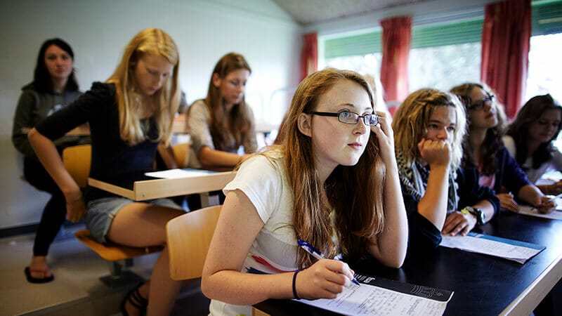 A classroom scene with several students seated at desks, attentively listening and taking notes. The room is well-lit, with windows and curtains in the background. The students appear focused on an unseen speaker or presentation.
