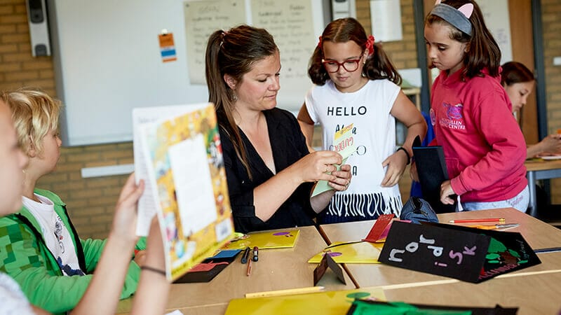 A teacher sits at a table in a classroom, engaging with several students. They are surrounded by colorful papers and craft supplies. The students, wearing casual clothing, watch attentively as the teacher demonstrates an activity.
