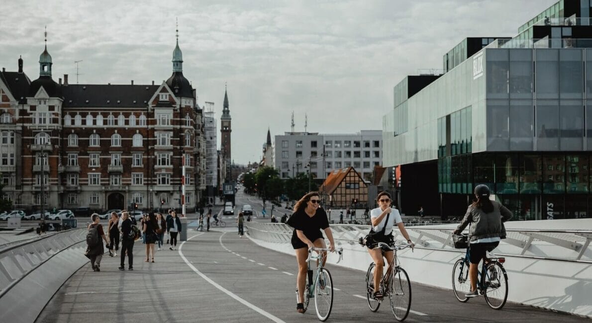 People are biking and walking on a modern bridge in an urban setting, with historical buildings and a modern glass structure in the background. The sky is overcast, adding a soft light to the scene.