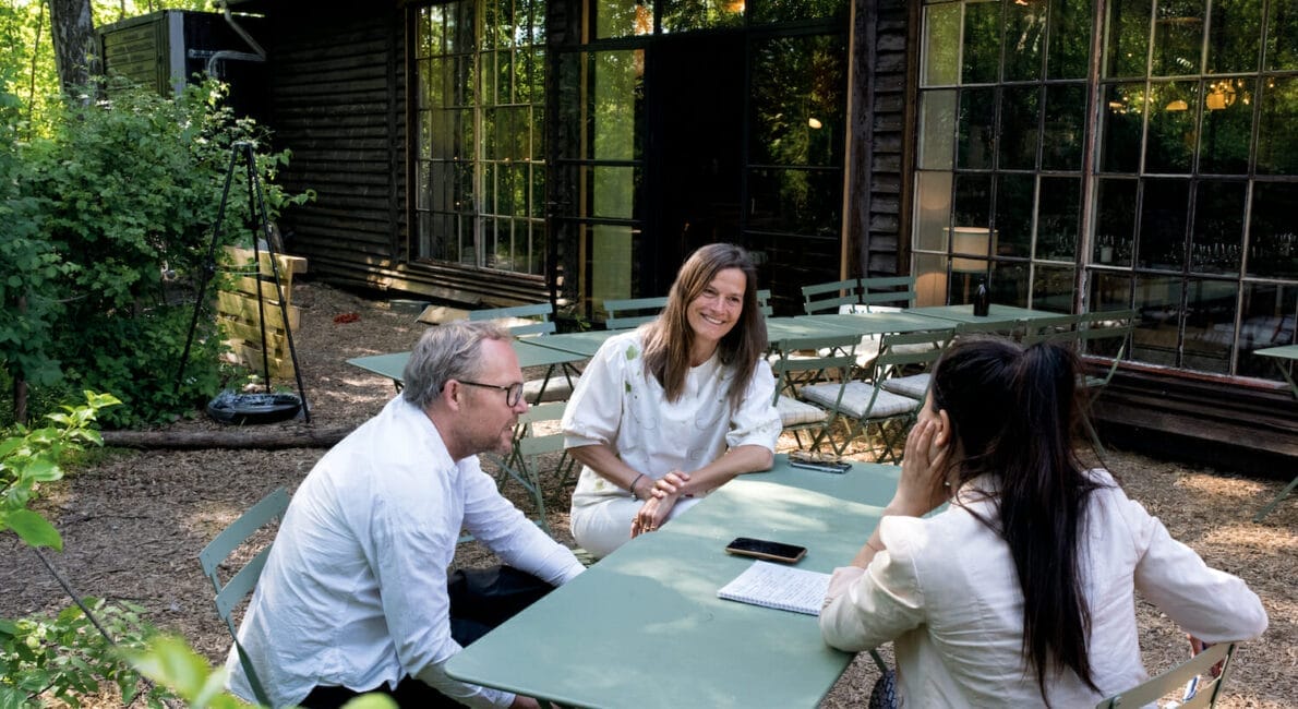 Tre personer sidder og taler ved et grønt bord udendørs. Indstillingen er skovklædt med en bygning med store vinduer i baggrunden. En person holder papirer, og en telefon ligger på bordet. Alle er afslappet klædt og fremstår i godt humør.