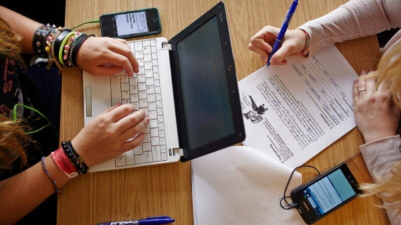 To personer arbejder sammen ved et træbord. Den ene skriver på en lille bærbar, mens den anden skriver på et udskrevet dokument. Begge har smartphones i nærheden, og flere blå kuglepenne ligger spredt på bordet.