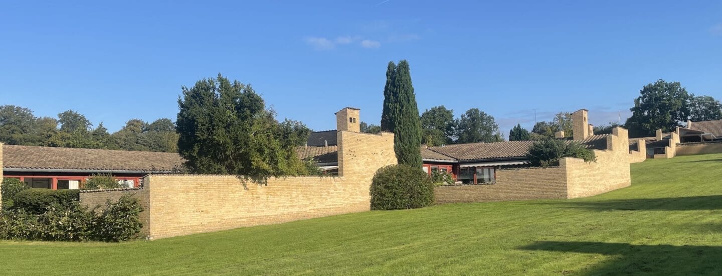 En naturskøn udsigt over en bygning med murstensvægge og et skrånende, græsklædt landskab under en klar blå himmel. Træer omgiver strukturen og tilføjer grønt til omgivelserne.