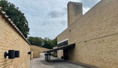 Stien flankeret af Fredensborghusene-inspirerede lysebrune murstensbygninger guider dig til et højt, smalt murstenstårn. Disse moderne, minimalistiske strukturer, fremhævet med slanke sorte væglamper, er sat på baggrund af træer under en overskyet himmel.