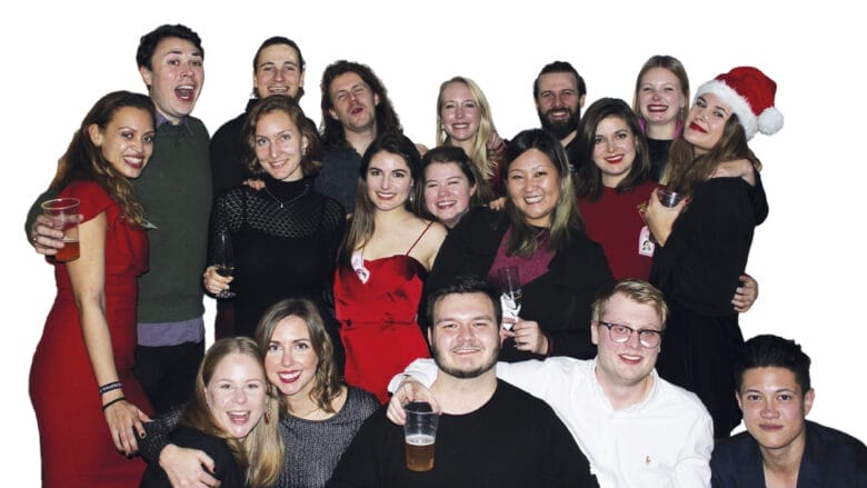 A cheerful group of people, possibly Danes, are posing together with smiles and holding drinks. One person dons a Santa hat, hinting at a festive or holiday gathering. A white background highlights the joyous scene in the image.