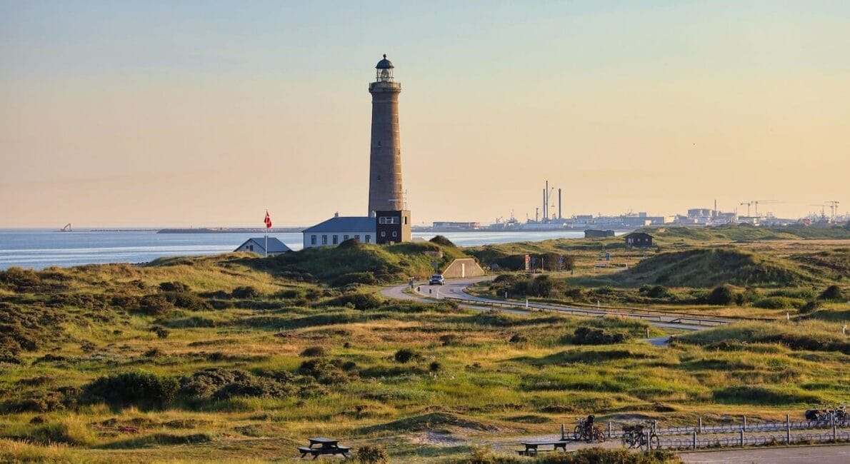 A tall lighthouse stands on a grassy coastal landscape during sunset. A winding road leads to the lighthouse, with a few scattered buildings and a flag nearby. The sky is clear, and the sea is visible in the background.