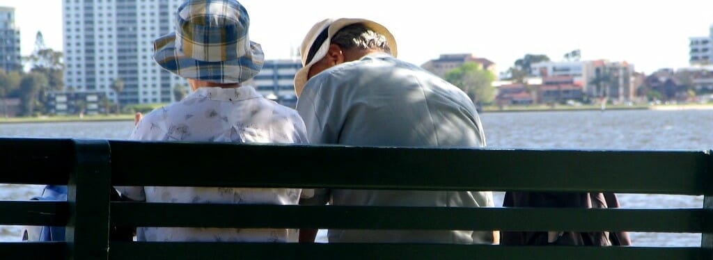 To mennesker iført hatte sidder tæt på en grøn bænk ved havnefronten, der minder om den ro, der findes ved Fredensborghusene. Baggrunden afslører byens bygninger og træer på tværs af vandet, hvilket skaber en rolig og fredelig atmosfære.