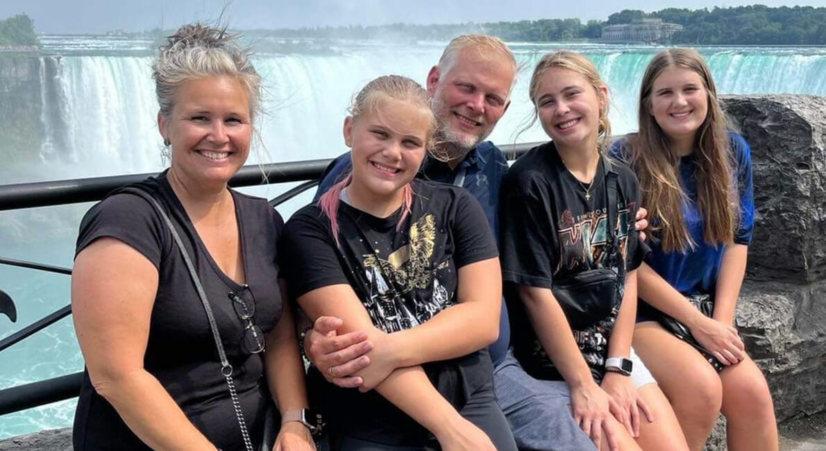 En familie på fem sidder på et rækværk foran et vandfald, sandsynligvis Niagara Falls. Gruppen omfatter to voksne og tre unge piger, der alle smiler til kameraet. De er afslappet klædt og ser ud til at nyde en solskinsdag.