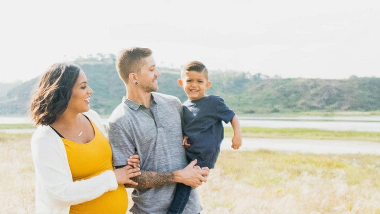 En gravid kvinde og en mand står udendørs på en græsmark. Manden holder et smilende barn i sine arme. Kvinden har en gul top og hvid cardigan på, manden en grå skjorte og barnet en mørkeblå skjorte. En sø og bakker er synlige i baggrunden.