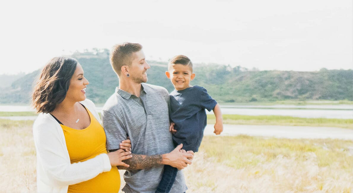 En gravid kvinde og en mand står udendørs på en græsmark. Manden holder et smilende barn i sine arme. Kvinden har en gul top og hvid cardigan på, manden en grå skjorte og barnet en mørkeblå skjorte. En sø og bakker er synlige i baggrunden.