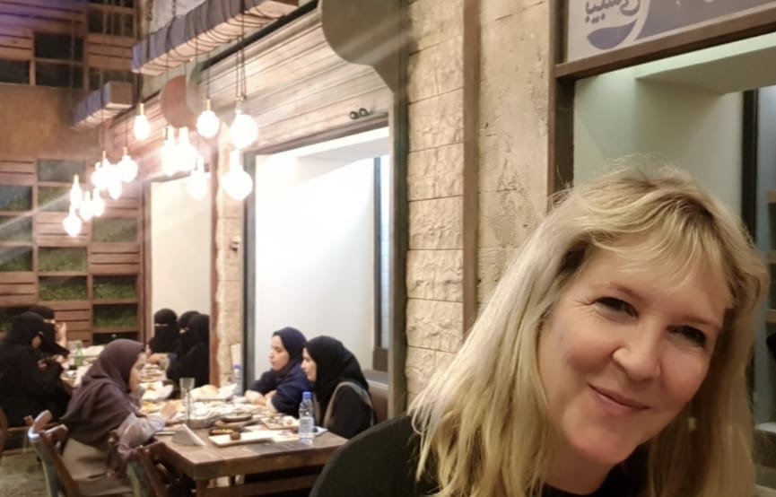 En kvinde med blond hår smiler til kameraet i en smart Dubai-restaurant. Bag hende sidder en gruppe mennesker ved et bord, dybt engageret i samtalen. Indstillingens varme belysning og træindretning skaber en indbydende atmosfære.