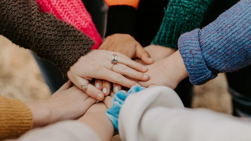 En gruppe mennesker stak deres hænder sammen i et show af sammenhold og teamwork. Klædt i farverige trøjer og et par ringe repræsenterer de samarbejde og solidaritet. Denne gestus afspejler danskernes globale ånd og fremmer forbindelse ud over grænser.