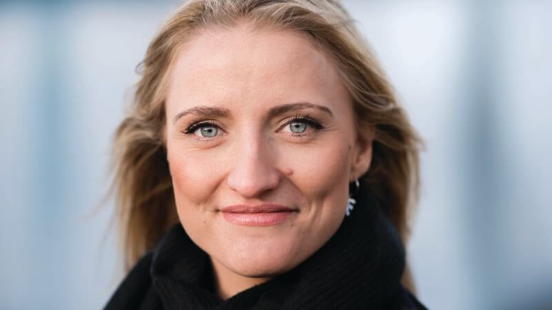 En person med langt blond hår og blå øjne smiler blidt, mens han stirrer direkte på kameraet. De er iført et sort tørklæde og øreringe, der udstråler en følelse af elegance mod en ufokuseret lys baggrund, der afspejler et subtilt strejf af kultur.