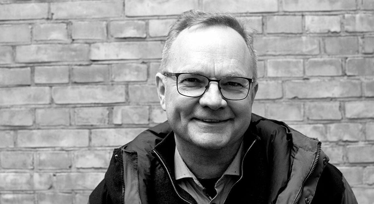 Sort/hvid billede af en smilende mand iført briller og en jakke med hætte. Han står foran en struktureret murstensvæg.