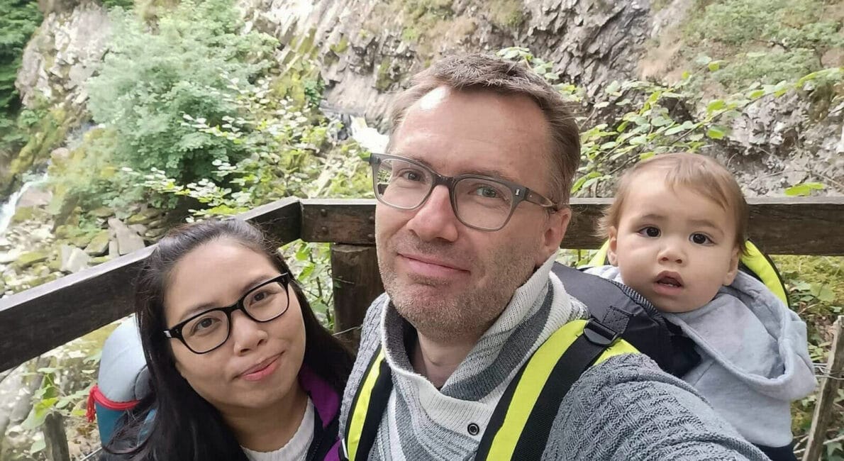En mand og en kvinde smiler til kameraet, mens de står på en træplatform foran et klippelandskab med grønne områder. Manden bærer en baby i en rygsækholder. Baggrunden har et vandfald og træer.