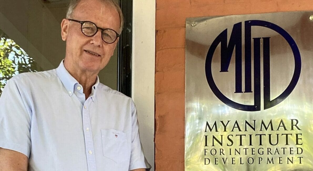 En person i en kortærmet skjorte med knapper står ved siden af et skilt til Myanmar Institute for Integrated Development. Skiltet har instituttets logo og hjemmeside. De er i et skyggefuldt udendørs område med en murstensvæg.