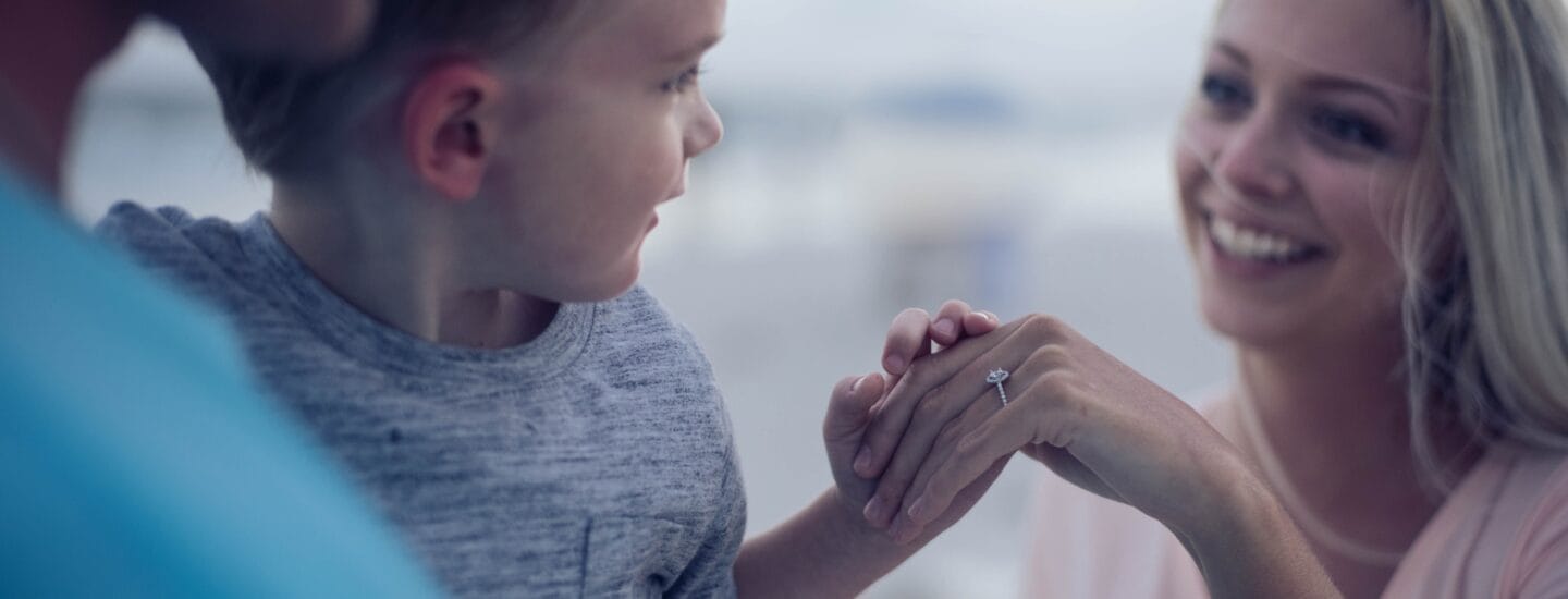 En kvinde med langt blondt hår smiler til en ung dreng i en grå skjorte, der holder hendes hånd, mens en mand i en blå skjorte står ved siden af dem. Den overskyede himmel antyder udendørs omgivelser, og fanger et øjebliks "familiesammenføring" under den store vidde.