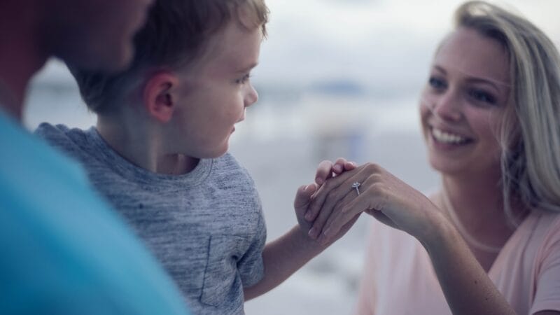 A woman with long blonde hair shows a ring on her hand to a smiling young boy and a man in a blue shirt. They are outdoors with a blurred, light-colored background.