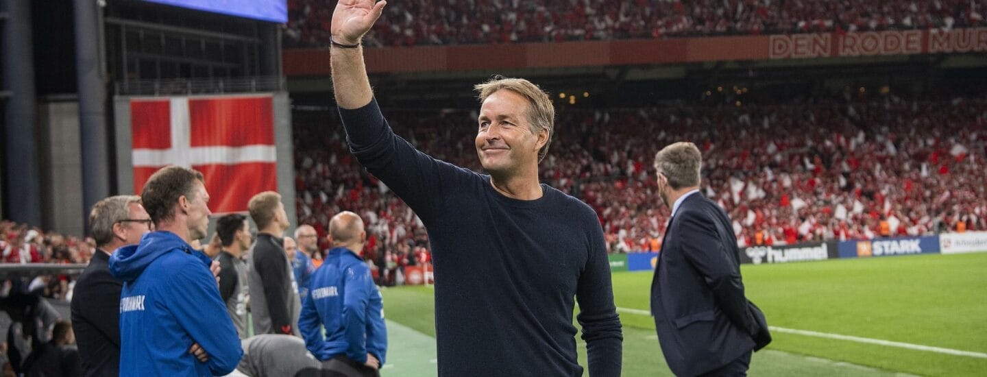 En mand i en sort sweater vinker til et jublende publikum på et stadion. Han står på sidelinjen af en fodboldbane, med trænere og holdpersonale i nærheden. Publikum holder røde flag, hvilket indikerer støtte til hjemmeholdet.