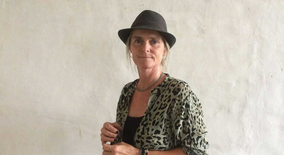 Person iført en top med leopardprint og en grå fedora-hat står op ad en almindelig lys væg. De har kort hår og holder et par briller i den ene hånd, med et neutralt udtryk.
