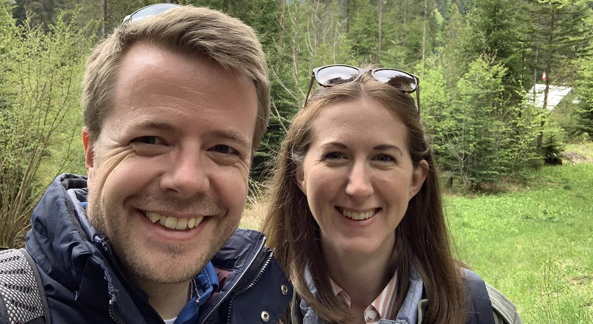 En smilende mand og kvinde tager en selfie udendørs i en frodig grøn skov. Begge er iført jakker, og kvinden har solbriller hvilende på hovedet. Baggrunden har træer og en antydning af et telt.