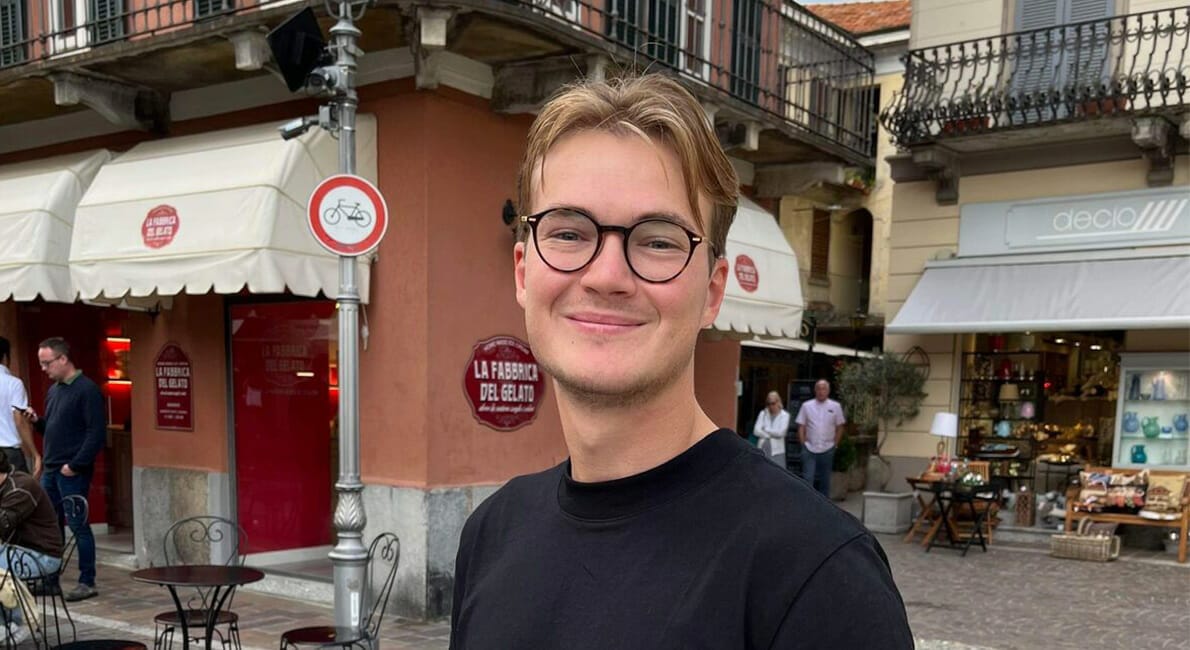 En smilende person med briller står på en brostensbelagt gade foran en café. Caféen har udendørs siddepladser og markiser. I nærheden er der butikker og et cykelforbud. Personen er iført en sort skjorte.