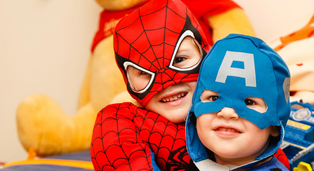 To små børn klædt i superheltekostumer. Den ene bærer et Spider-Man-dragt, mens den anden er i Captain America-outfit. De smiler og krammer, med en stor udstoppet legetøjsbjørn i baggrunden.