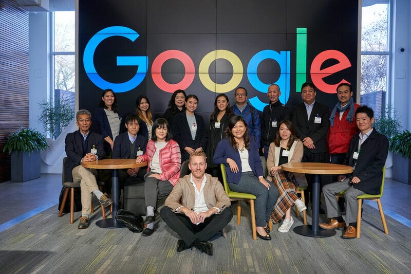 En gruppe mennesker poserer til et billede foran et stort Google-logo. Nogle sidder ved borde, mens andre står bag dem. De er i et moderne interiør med planter og en skærm med logoet.