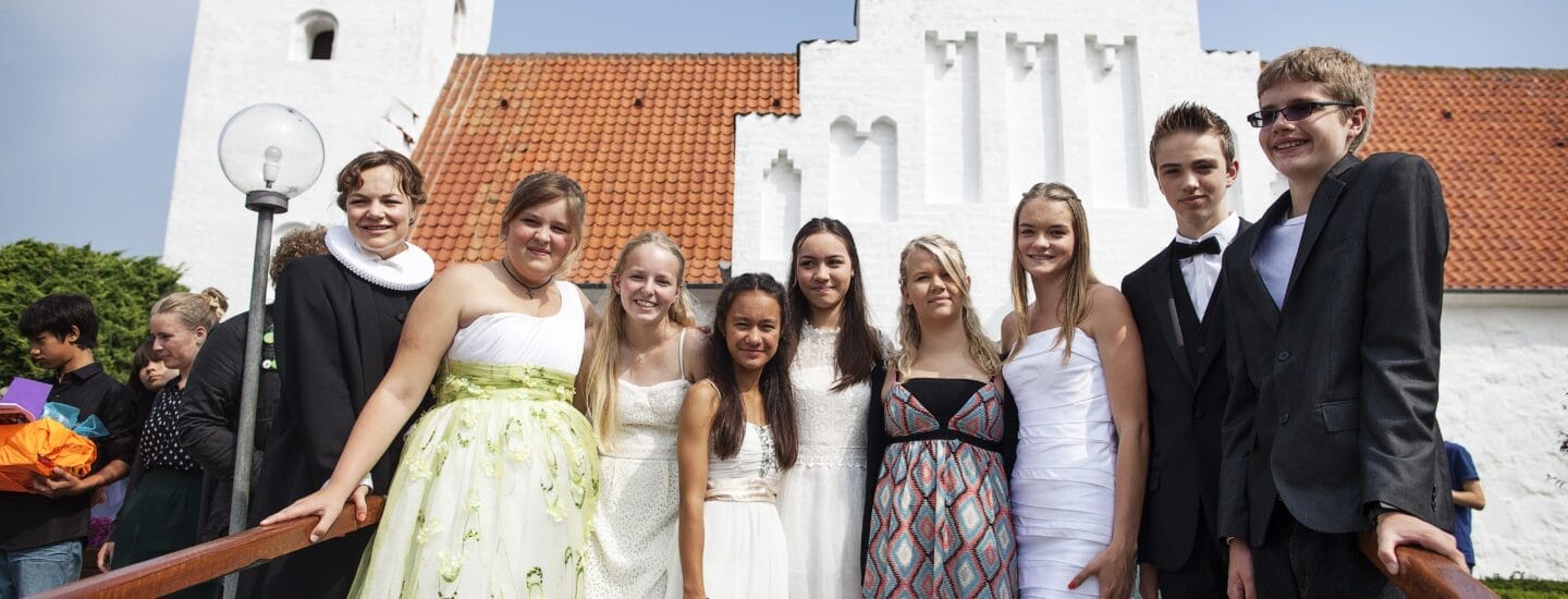 A group of young people dressed in formal attire stand together outside a white church with a red-tiled roof. They are smiling and posing for the photo on a sunny day.