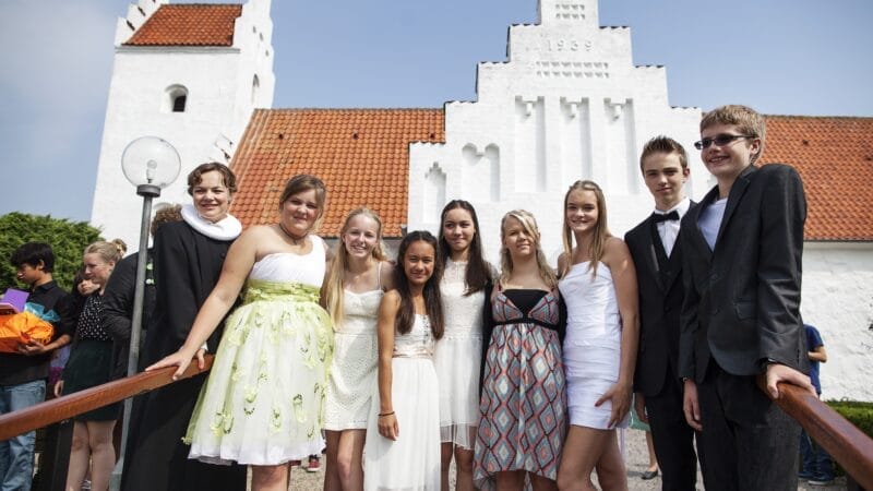 En gruppe unge fra Den Danske Sommerskole, klædt i højtidlig påklædning, poserer foran en hvid kirke med trappegavl og rødt tag. Himlen er klar, da hver person smiler til kameraet.