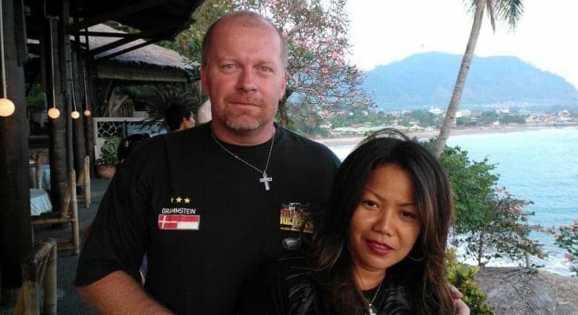 En mand og kvinde står sammen på et trædæk med udsigt over havet. Manden har en sort skjorte med logoer på, og kvinden har langt mørkt hår. Der er et træ og et bjerg i baggrunden under en klar himmel.