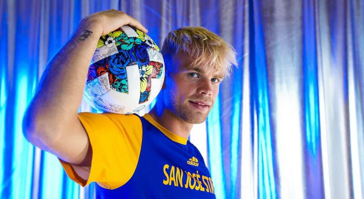 En fodboldspiller poserer selvsikkert i en blå og gul "San Jose State"-trøje og holder en fodbold på sin skulder. Baggrunden har metalliske blå og sølv gardiner, der skaber en dynamisk atmosfære.