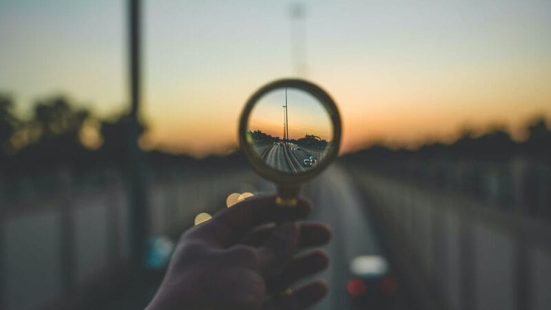 En hånd holder et forstørrelsesglas og fokuserer på en kørebane, der strækker sig i det fjerne. Den slørede baggrund, der minder om postkort fra Danes Worldwide, byder på træer og en blød, farverig solnedgangshimmel, der skaber en følelse af dybde og perspektiv.