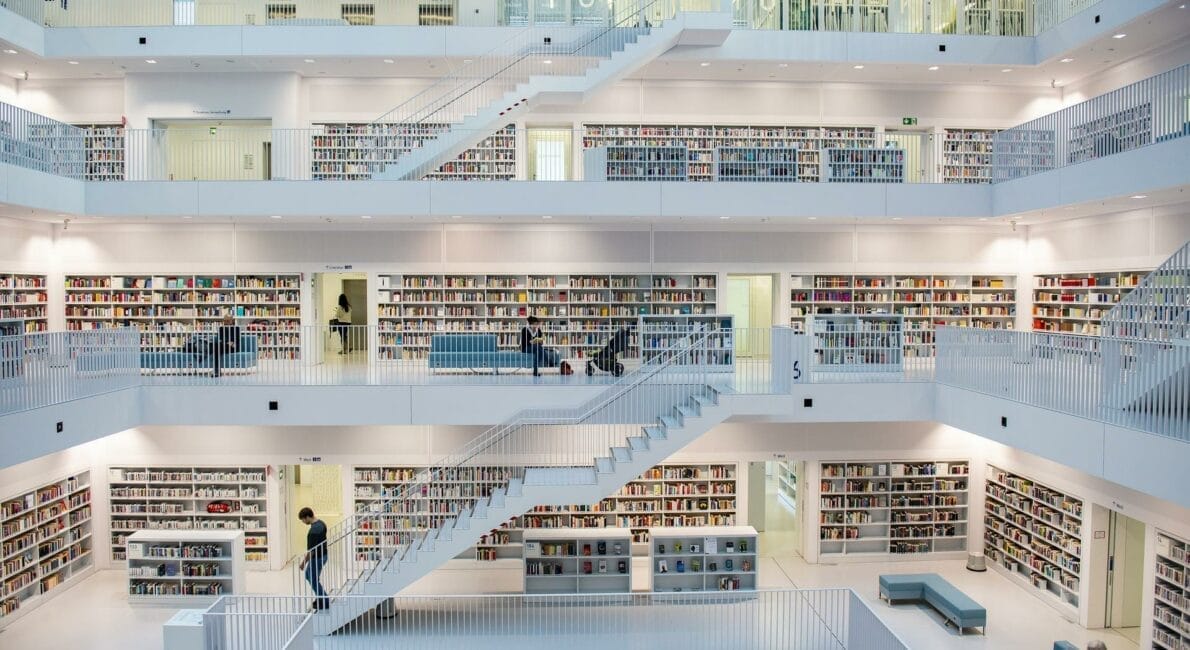 Et rummeligt, moderne bibliotek med flere niveauer af hvide balkoner foret med bogreoler. Folk læser og går mellem hylderne. Et centralt åbent område er synligt med trapper, der forbinder etagerne.