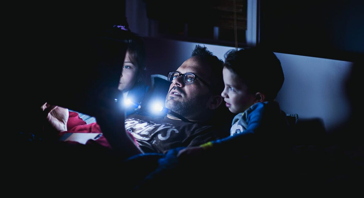 En mand med briller sidder i et svagt oplyst rum med to børn og læser eller ser noget på en tablet. Lyset fra skærmen oplyser deres ansigter og skaber en hyggelig atmosfære, når de hviler mod puder.