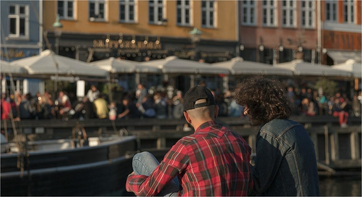 To personer sidder ved en havnefront med en båd i forgrunden. Den ene bærer en rød plaid skjorte og kasket, den anden en denimjakke. En travl udendørs café med paraplyer og mennesker er i baggrunden, med farverige bygninger langs scenen.