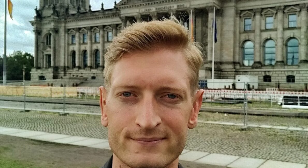 En person med lyst hår iført sort jakke tager en selfie foran rigsdagsbygningen i Berlin. Den historiske struktur har en stor glaskuppel og flag. Himlen er overskyet.