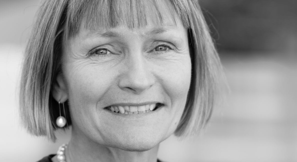 Et sort/hvidt portræt af en person med skulderlangt hår og en perlehalskæde, der smiler til kameraet. Baggrunden er blødt sløret.