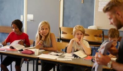 Fire børn sidder ved et bord i et klasseværelse og engagerer sig i deres åbne bøger. En lærer står i nærheden og holder papirer. Klasselokalet har blå vægge og en stor korkplade i baggrunden.