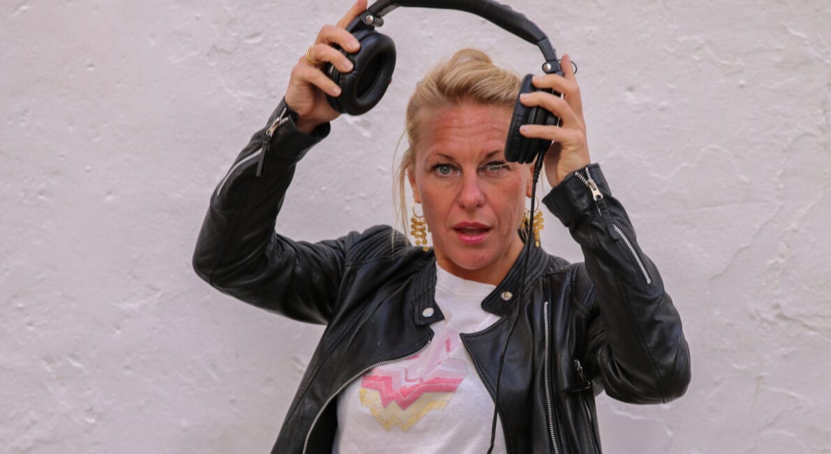 En digital nomade iført en sort læderjakke og store øreringe justerer et par hovedtelefoner på hovedet. De står på en tekstureret hvid baggrund, og ser direkte på kameraet og inkarnerer essensen af moderne, mobil kreativitet.