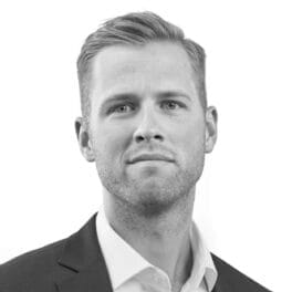 Et gråtonebillede af en person med kort, lyst hår, iført en mørk jakkesæt og en skjorte med hvid krave, og kigger direkte på kameraet mod en almindelig hvid baggrund.