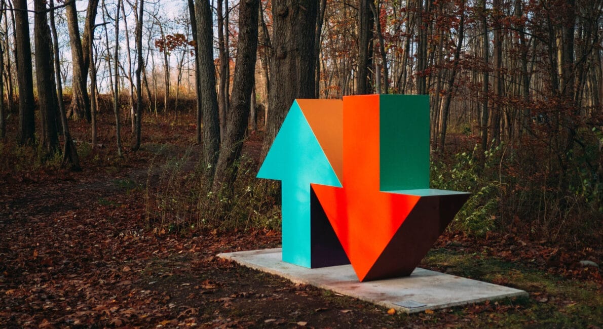 En stor, farverig skulptur i form af to pile, den ene peger op i turkis og den anden ned i orange, står på mystisk vis på en piedestal midt i en skov af nøgne træer og spredte efterårsblade.