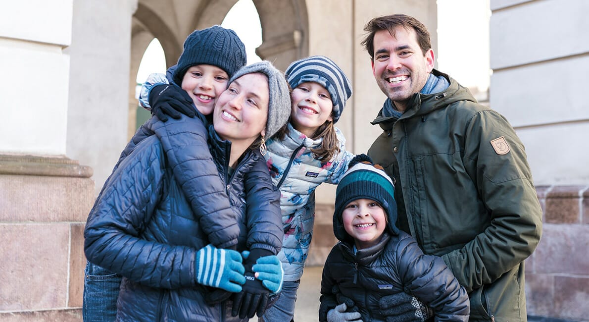 En glad familie på fem, iført vinterjakker og huer, smiler udendørs. De står tæt sammen foran en stenbygning med buer og ser glade og hyggelige ud i det kølige vejr.