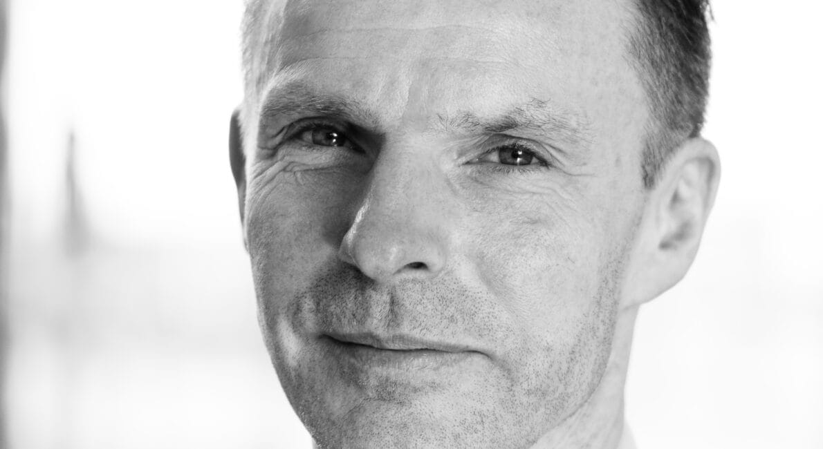 Sort og hvidt portræt af en mand iført et jakkesæt med en hvid skjorte. Han har kort hår og kigger direkte på kameraet med et lille smil. Baggrunden er blødt sløret.