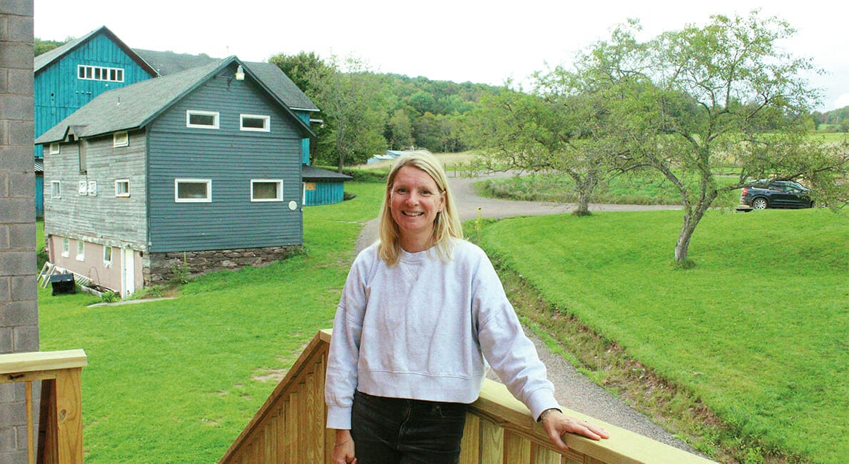 En person med langt hår står på et trædæk iført en let sweater og sorte bukser. Bag dem er en grøn græsplæne, et par træer, en grussti og et blåt hus. En bil er parkeret i baggrunden under en overskyet himmel.