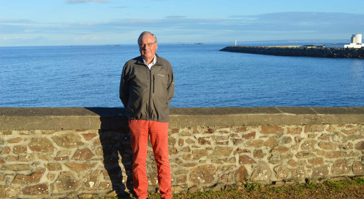 En person står foran en stenmur med udsigt over et roligt blåt hav. De er iført en grå jakke og røde bukser. Himlen er klar med enkelte skyer, og en fjern mole eller bølgebryder strækker sig ud i vandet.