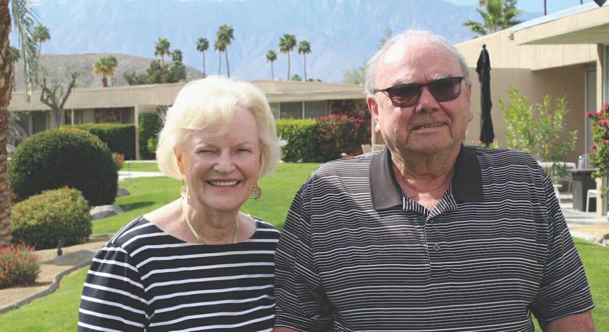 Et ældre par smilende udendørs med palmer og bjerge i baggrunden. Begge er iført stribede skjorter, og manden har solbriller på. De står i et haveområde med velholdt græs og buske.