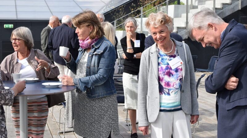 A group of people are gathered outdoors, chatting and holding drinks. Some are standing at a table while others are mingling. The setting is bright with a glass structure in the background, and everyone appears engaged in conversation.