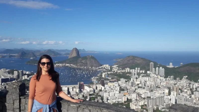 A woman with long hair, wearing sunglasses and an orange shirt, stands smiling in front of a scenic city by the sea. A network of boats dots the bay beneath the famous mountain, all set against a clear blue sky.