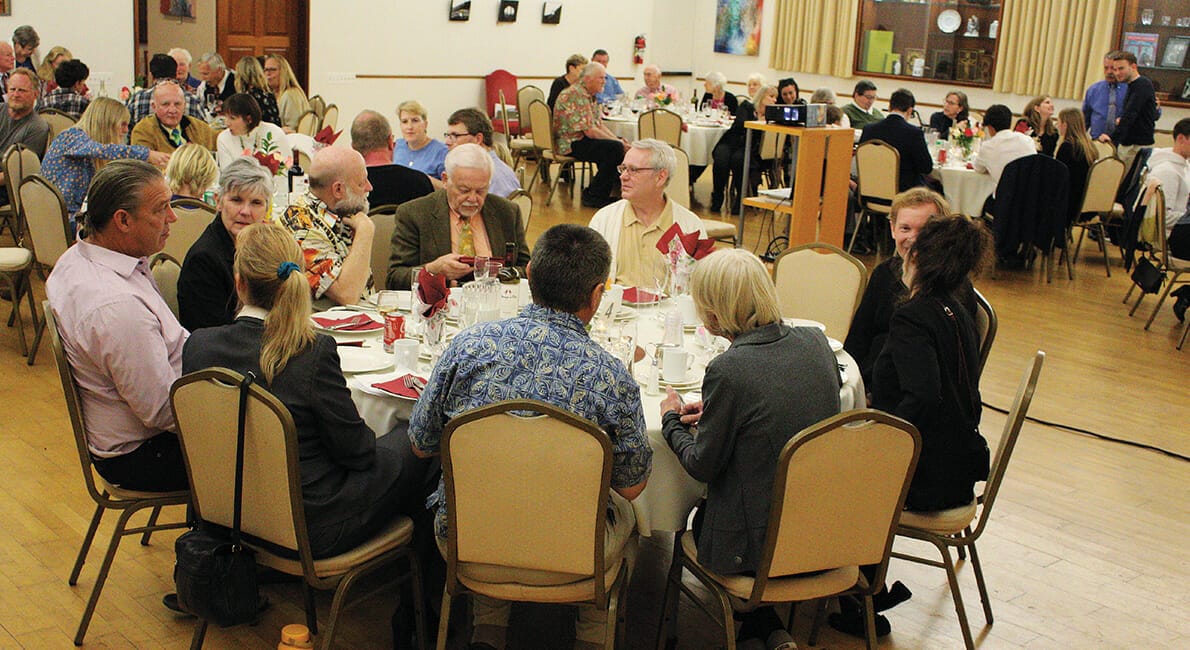 En stor gruppe mennesker sad omkring cirkulære borde i en festsal og var engageret i samtale. Bordene er dækket med hvide duge, røde servietter og glas. Deltagerne er klædt i en blanding af afslappet og formel påklædning.