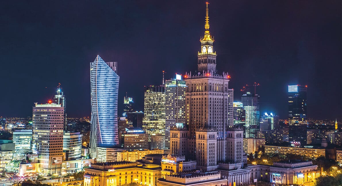 En natudsigt over Warszawas bybillede med det oplyste Kultur- og Videnskabspalads. Omgivende skyskrabere lyser af lys og fremhæver moderne arkitektur mod den mørke himmel.